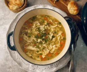 A steaming bowl of homemade rotisserie chicken noodle soup garnished with fresh herbs, featuring tender chicken, hearty noodles, and colorful vegetables.